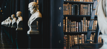 Corredor de uma biblioteca, com várias estantes cheias de livros, no início de cada estante há um busto de uma estátua